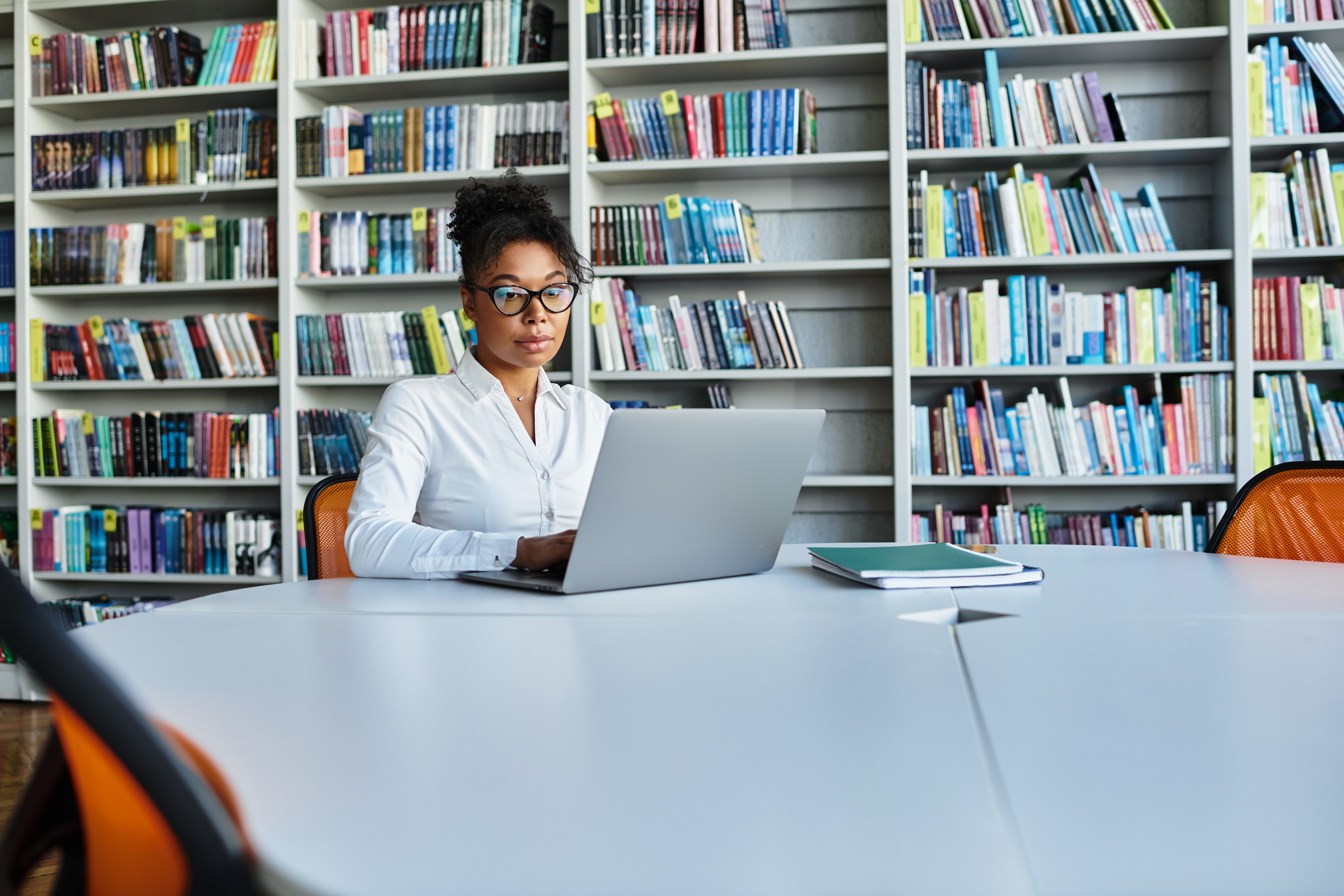 A young woman with glasses and a white shirt sitting at a large library table, working on a laptop with a backdrop of tall, colorful bookshelves.