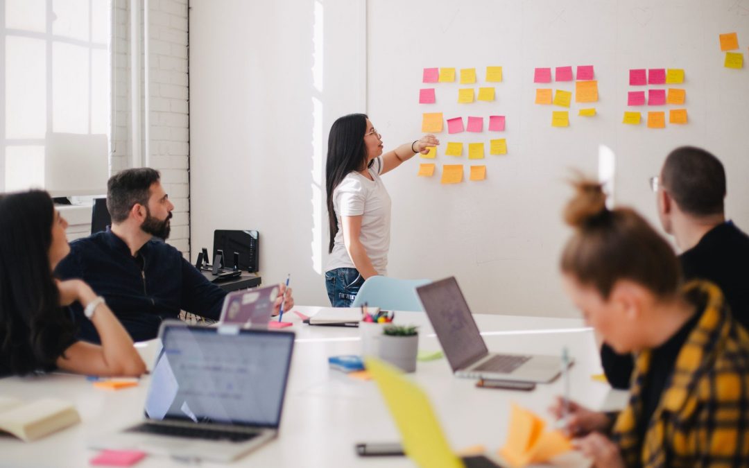 A group of people in an office or classroom setting. A woman is standing at a whiteboard covered in colorful sticky notes, pointing to one of them while speaking. Other individuals are seated around a table with laptops and notebooks, looking at the whiteboard.