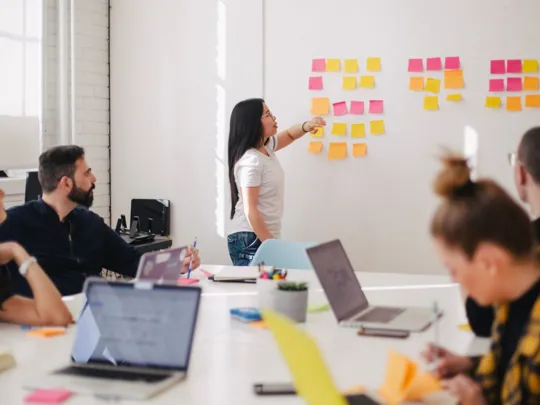 A group of people in an office or classroom setting. A woman is standing at a whiteboard covered in colorful sticky notes, pointing to one of them while speaking. Other individuals are seated around a table with laptops and notebooks, looking at the whiteboard.