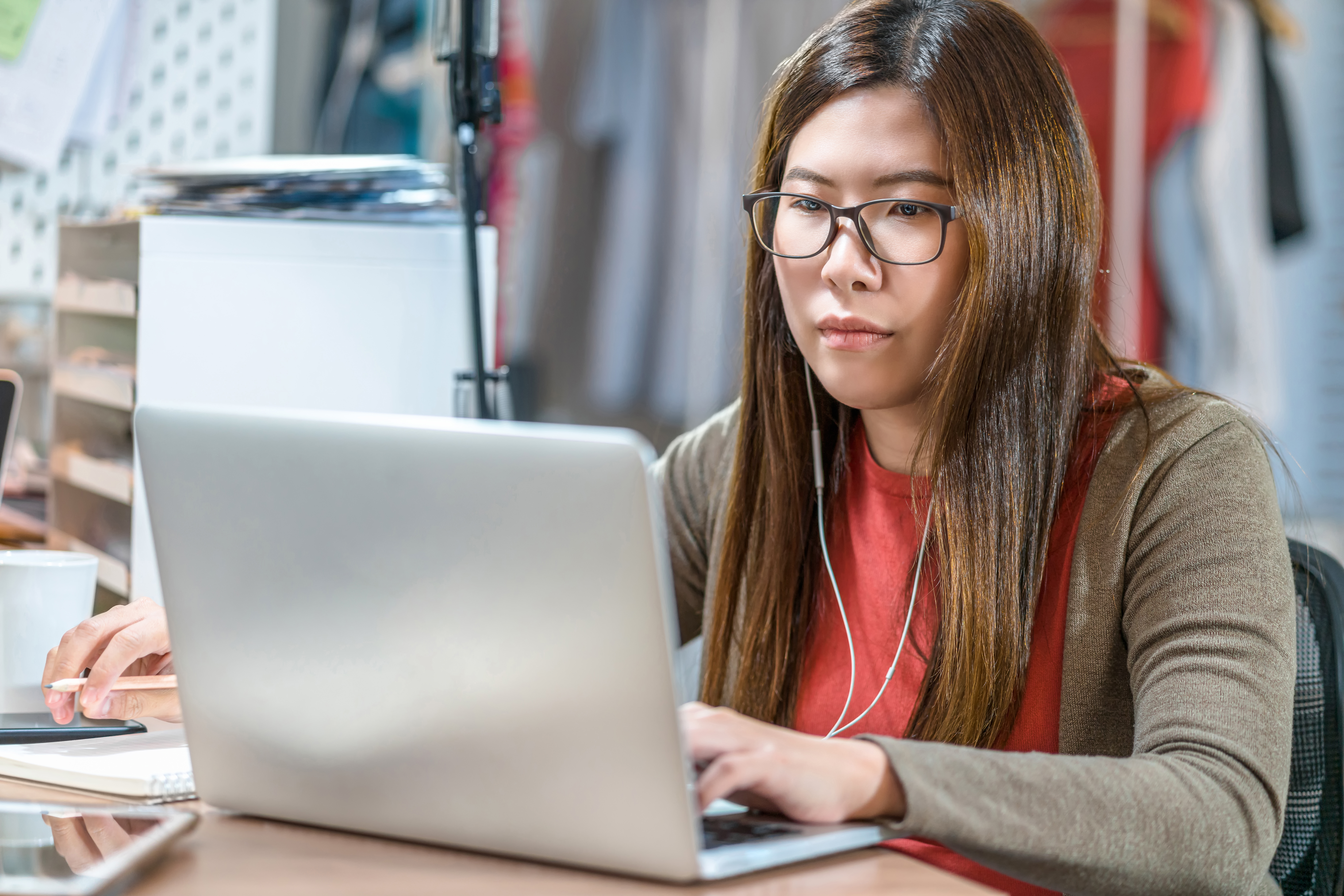 A young woman wearing glasses and earbuds intently works on a silver laptop at a desk, representing focused online learning or remote work.