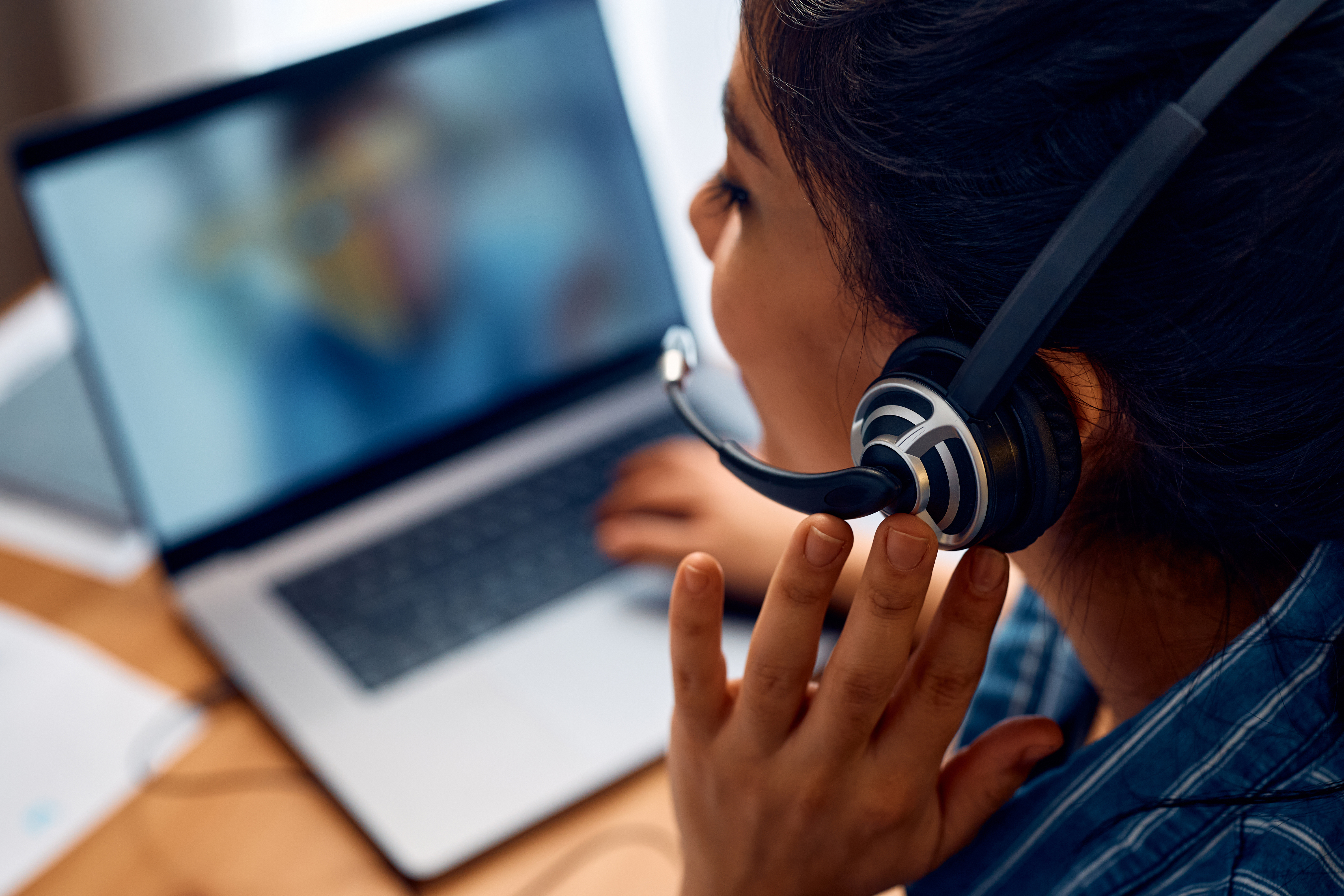 A woman wearing a headset with a microphone is talking on a video call on her laptop. 