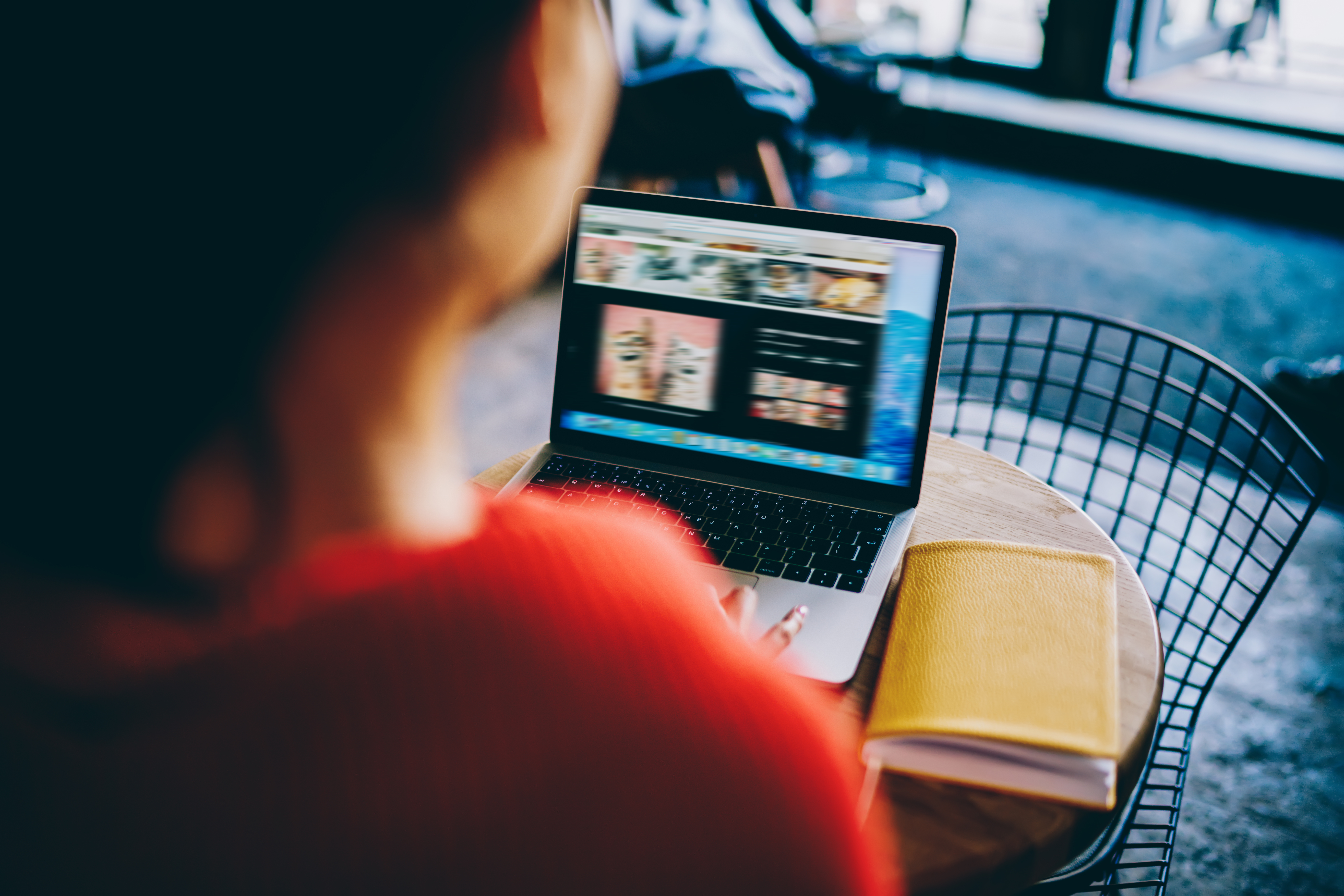 A person in a red shirt working on a laptop displaying a website photo grid, with a yellow notebook nearby, set in a trendy cafe.