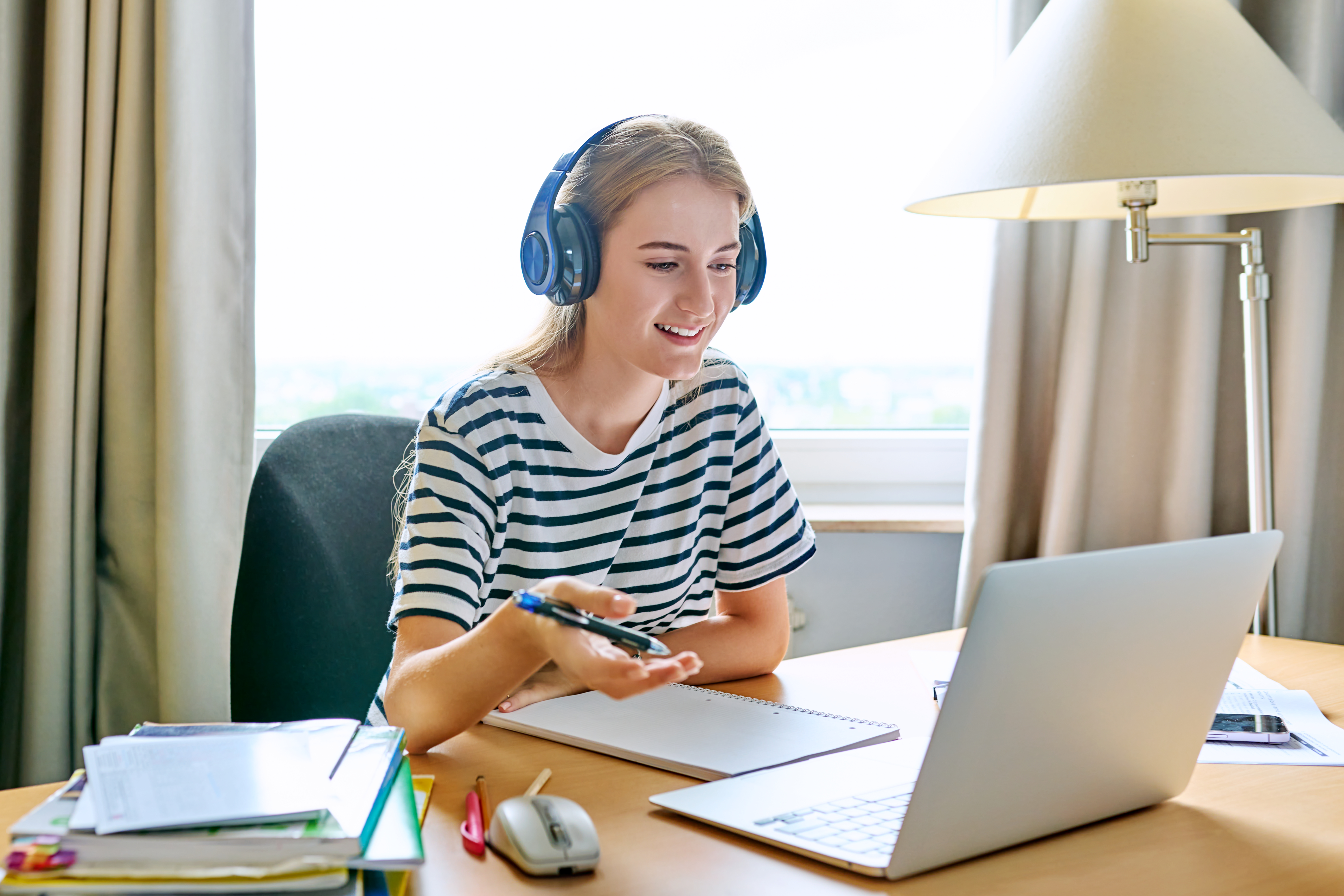 a young female student wearing headphones and a striped shirt, smiling while studying at a laptop for an online class at a desk.
