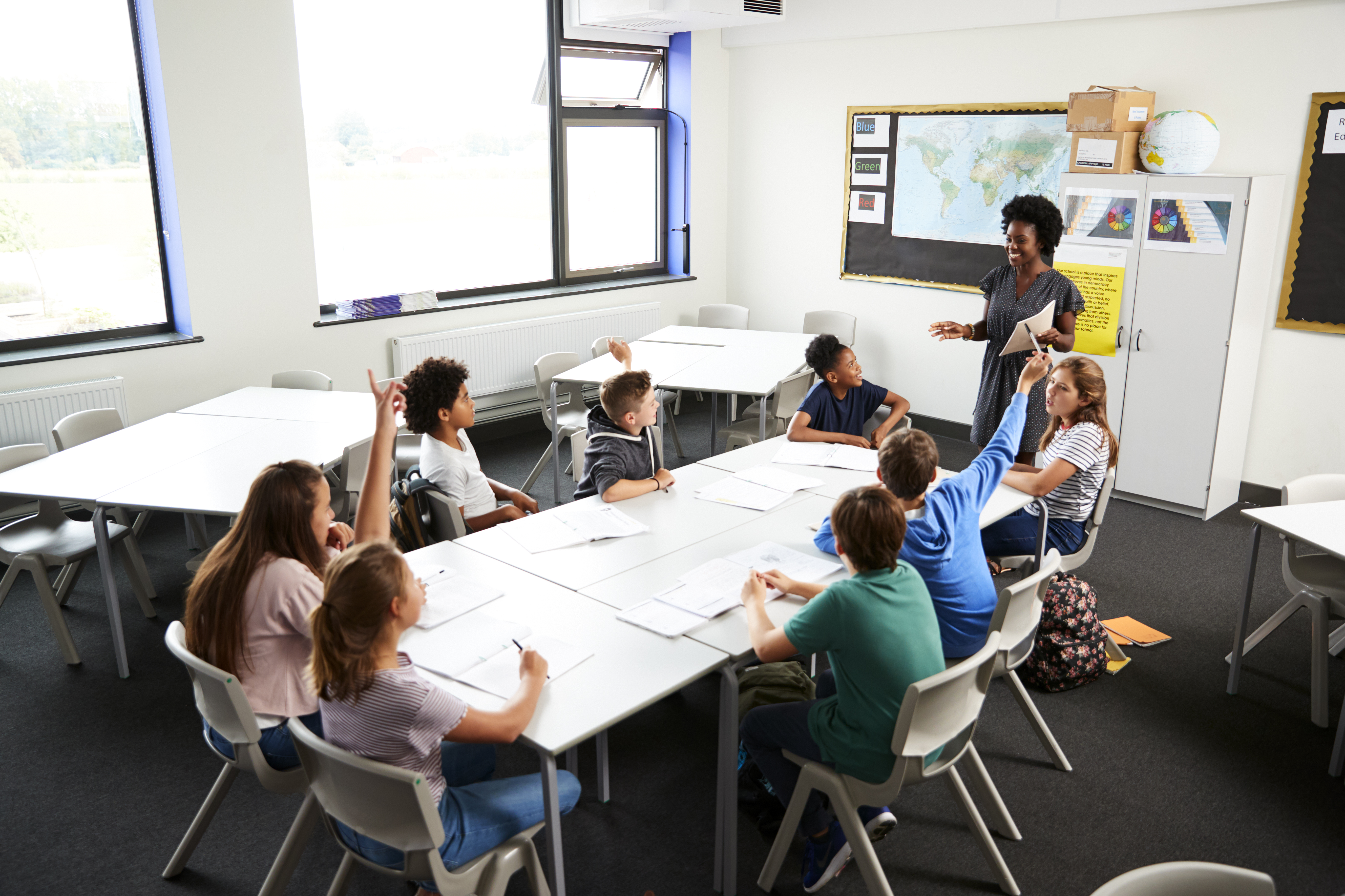 Teacher standing up in front of a class of students sat down with their hands up.