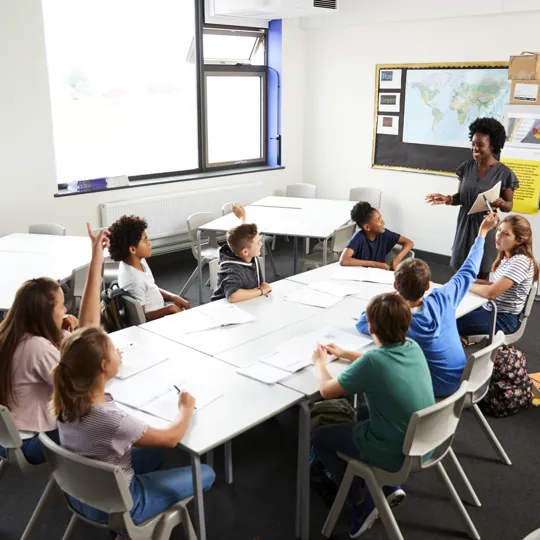 Teacher standing up in front of a class of students sat down with their hands up.