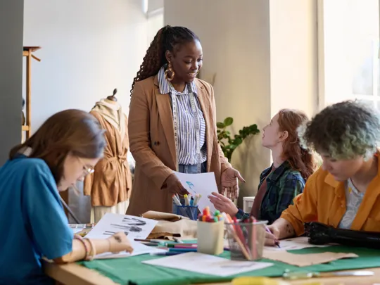 A friendly female instructor with a measuring tape around her neck smiles while presenting a design paper to a group of students gathered around a table with art supplies. They are working on a fashion or design project in a well-lit studio or classroom.