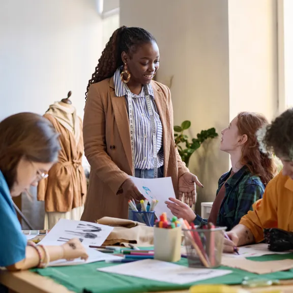 A friendly female instructor with a measuring tape around her neck smiles while presenting a design paper to a group of students gathered around a table with art supplies. They are working on a fashion or design project in a well-lit studio or classroom.